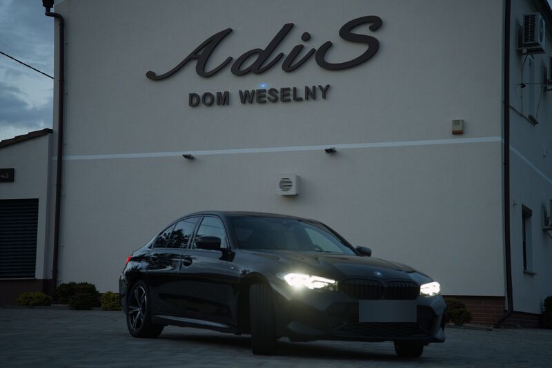 Ciemny sedan BMW przed szyldem Domu Weselnego ADIS